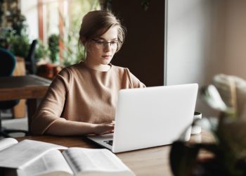 Work, lifestyle and business concept. Good-looking focused european female in trendy glasses sitting in cafe near laptop, working on notebook, surrounded with books, making notes for lesson with kids.