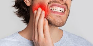 Tooth and gum inflammation. Guy suffers from pain in his mouth and presses hand to red sore spot, panorama, cropped, studio shot