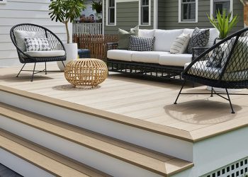 A low-to-the-ground composite deck is built into a small alcove on the back of a home. The deck features two wide stairs that lead to a stone patio. The boards are a warm tan and white PVC trim is used in fascia and stair riser applications. Furniture with black woven frames and white cushions lends a Scandinavian feel to the space. Touches of greenery in pots add color and texture to the space. The composite deck is made of Weathered Teak Multi-Width Decking from the TimberTech AZEK Vintage Collection.